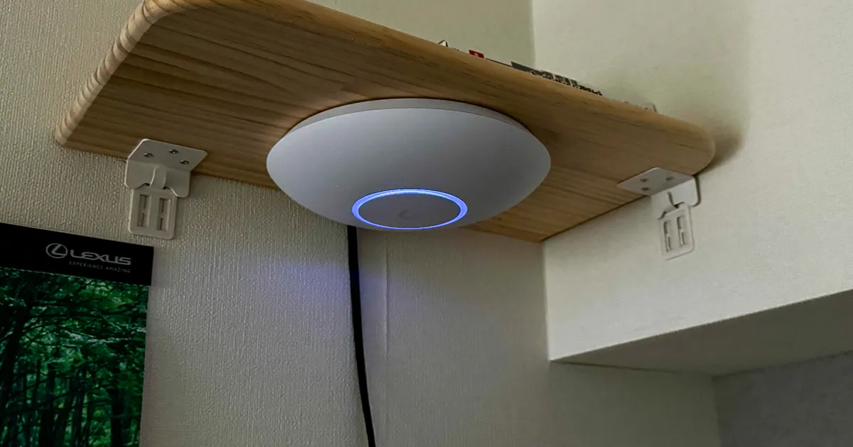 壁美人(ホッチキス固定)で付けた木製棚の下面に U7 Pro を下向きに設置。青い LED リングが点灯し、PoE ケーブルが壁を伝って下りている